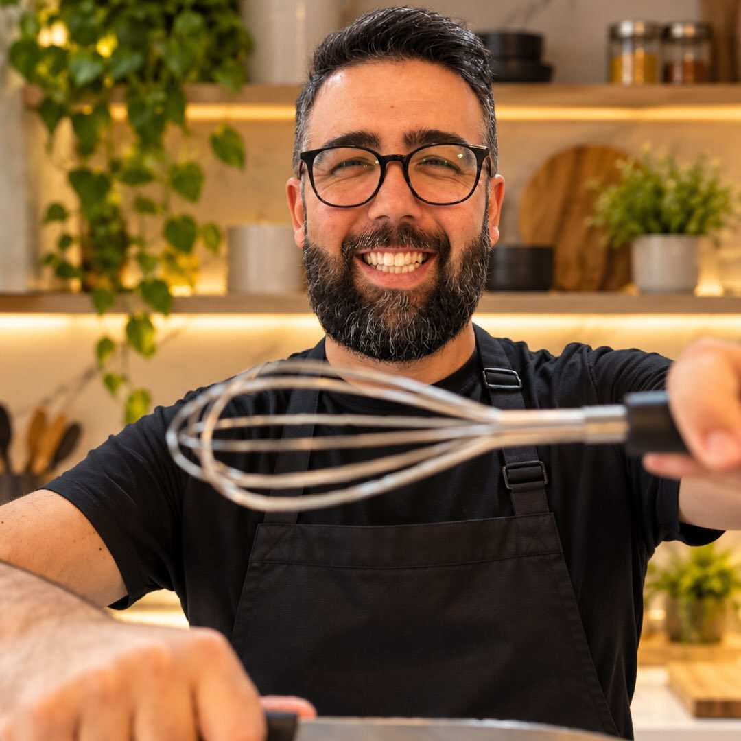 Chef Rafael Marcelino sorrindo em sua cozinha, com avental preto e um fouet